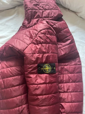 Vinröd pufferjacka från Stone Island - Säljer en vinröd pufferjacka från Stone Island med huva och klassisk badge på ärmen. Jackan är quiltad och har dragkedja framtill. Materialet är glansigt och syntetiskt, perfekt för dig som gillar streetwear och vill sticka ut med en snygg färg.