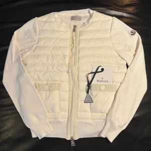 Moncler Cardigan  - Snygg vit cardigan jacka från Moncler med quiltad front och stickade ärmar. Jackan har dragkedja, rund hals och två fickor framtill. Moncler-logga på ärmen och ribbade muddar. Perfekt för dig som gillar stilren design och exklusiva märken.