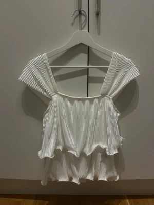 Vit plisserad linnetopp med volang - Säljer en vit linnetopp med breda plisserade axelband och dubbla lager volanger. Toppen har en luftig och flowy känsla med snygga veckade detaljer som ger extra stilpoäng. Perfekt för dig som gillar en unik och trendig look.