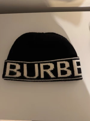 Burberry mössa - Säljer en svart mössa från Burberry med bred vit logotyp runt hela kanten. Klassisk och stilren design som passar till det mesta. Mössan är stickad och har en mjuk känsla.