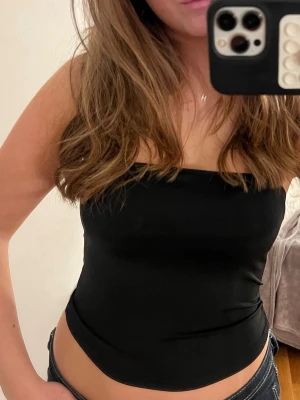Svart bandeau tubtopp - Säljer en svart bandeau tubtopp med tight passform. Toppen är axelbandslös och sitter snyggt mot kroppen, perfekt till jeans eller kjol. Enkel och stilren design som är lätt att matcha med olika outfits.