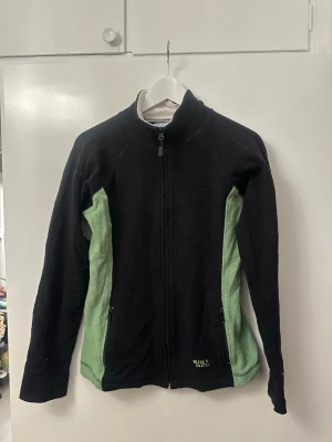 Svart och grön fleecejacka från Bik Bok - Mysig fleecejacka från Bik Bok i svart med ljusgröna paneler längs sidorna. Jackan har hel dragkedja framtill, två fickor med dragkedja och hög krage. Perfekt för lager-på-lager och chill dagar. Broderad logga på ena fickan.