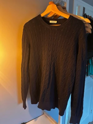 Marinblå kabelstickad tröja J.Lindeberg - Kabelstickad marinblå tröja från J.Lindeberg med rund hals och diskret logodetalj i metall. Tröjan har lång ärm och ribbade muddar vid hals, ärmslut och nederkant. Perfekt för dig som gillar klassisk stil med en modern touch. Däremot passar den bättre till storlek M.