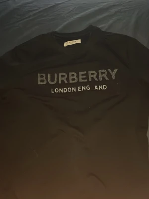 Burberry tröja  - Säljer en svart t-shirt från Burberry med stor logga och texten 'London England' broderat på bröstet. Klassisk rund hals och korta ärmar. Perfekt för dig som gillar stilrena designer och vill ha något från ett ikoniskt märke.
