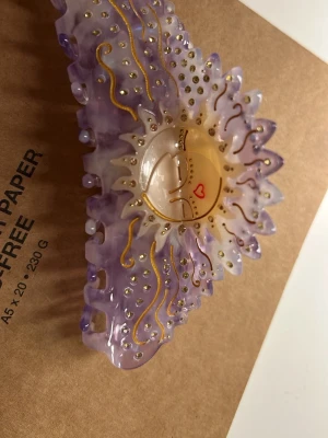 Ljuslila hårklämma från Maanesten - Unik hårklämma från Maanesten i ljuslila och beige med sol- och månansikte i mitten. Klämman har guldfärgade detaljer, små glittriga stenar och vågiga former. Perfekt accessoar för att lyfta din look med en magisk touch. Fick den i julklapp men den va inte ricktigt min stil , alldig andvänd !❣️🐻