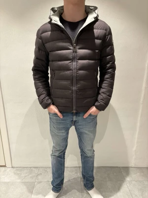 Colmar pufferjacka - Säljer en mörkgrå pufferjacka med huva och dragkedja framtill. Jackan har två sidofickor med dragkedja. Insidan av huvan är ljusgrå vilket ger en snygg kontrast. Perfekt för kyliga dagar. Loggan på jackan har lite defekter. Jackan är i storlek S