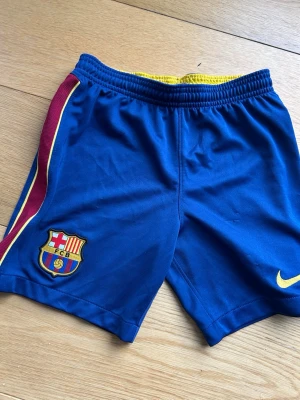 Barça shorts med Nike-logga - Snygga blå FC Barcelona shorts med gul Nike-logga och klubbmärke på ena benet. Röd och gul rand längs sidan ger extra stilpoäng. Perfekta för fotbollsträning eller häng med kompisarna. Elastisk midja för skön passform.