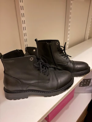 Svarta boots från Jack & Jones - Säljer ett par svarta boots från Jack & Jones med grov sula och snörning. Skorna har dragkedja på sidan för enkel på- och avtagning och är tillverkade i skinn. Perfekta för dig som gillar robust stil och vill ha något som funkar till många outfits. Ordinarie pris 1499kr.