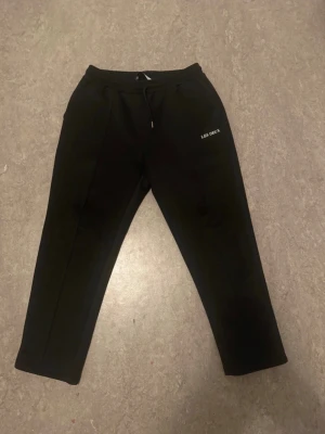 Svarta mjukisbyxor från Les Deux - Säljer ett par svarta mjukisbyxor från Les Deux med dragsko i midjan och diskret logga på benet. Byxorna har raka ben och sidofickor, perfekta för chill dagar eller när du vill vara bekväm men ändå se stilren ut.