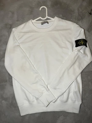 Vit sweatshirt från Stone Island - Vit sweatshirt från Stone Island med klassisk rund hals och ribbade muddar. Tröjan har lång ärm och den ikoniska Stone Island-patchen på vänster ärm. Materialet är mjuk bomull som ger en skön känsla. Perfekt för dig som gillar stilrena och enkla plagg med tydlig streetwear-vibe. Passar 160 och uppåt 