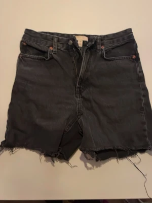 Svarta jeansshorts med rå kant - Säljer ett par svarta jeansshorts med rå, fransig kant och klassisk femficksdesign. Shortsen har knapp och dragkedja framtill samt nitar vid fickorna. Perfekta för dig som gillar en avslappnad och edgy stil.