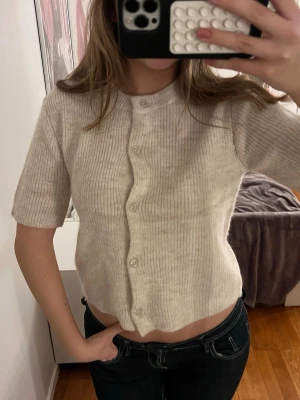 Beige stickad kofta från Gina Tricot - Superfin beige stickad kofta från Gina Tricot med kort ärm och ribbad struktur. Koftan har rund halsringning och knäpps med knappar framtill.  Passar S - M