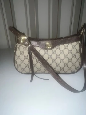 Beige och brun axelväska från Gucci - Säljer en snygg axelväska från Gucci med klassiskt GG-mönster i beige och brunt. Väskan har detaljer i brunt skinn, guldfärgad logga och dragkedja upptill. Insidan är ljus och mjuk, perfekt för dina essentials. Stilren och ikonisk design som lyfter din outfit.