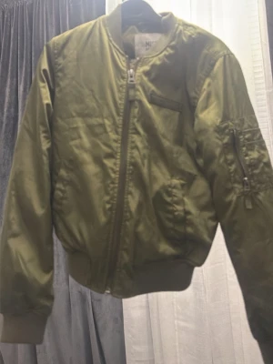 Olivgrön bomberjacka från Missguided - Snygg olivgrön bomberjacka från Missguided med klassisk passform. Jackan har dragkedja framtill, fickor på sidorna och en extra ficka med dragkedja på ärmen. Tillverkad i glansig polyester med ribbade muddar vid ärmslut och midja.