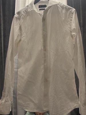 Vit skjorta från Riley - Klassisk vit skjorta från Riley med lång ärm och knappar framtill. Skjortan har en stilren krage och är tillverkad i ett tunt, mjukt bomullstyg som känns skönt mot huden. Perfekt för dig som gillar en clean och enkel look.