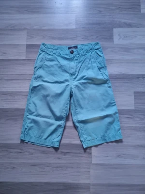 Turkosa shorts från H&M L.O.G.G. strl 164 - Säljer ett par turkosa shorts från H&M L.O.G.G. i storlek 164. De har normal passform, bälteshällor, knapp och dragkedja framtill samt fickor både fram och bak. Materialet är mjuk bomull, perfekta för varma dagar.
