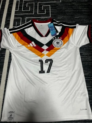 Vit Adidas fotbollströja Tyskland #17 - Säljer en vit Adidas fotbollströja för Tysklands landslag med nummer 17 och namnet Wirtz på ryggen. Tröjan har mönster i svart, rött och gult över bröstet, v-ringad krage med röd kant och broderat landslagsemblem. Tillverkad i polyester med ClimaCool-teknologi för snabb torkning.