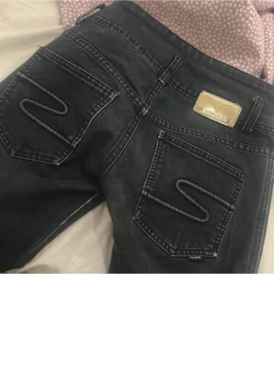 Svarta lågmidjade bootcut jeans - Y2K vibe  - Säljer ett par sjukt snygga lågmidjade jeans i mörkblå tvätt 🤍 Perfekta för Y2K / 2000-tal / vintage-stil. Jeansen sitter tight över höfter & rumpa och går lätt ut nertill (bootcut).  Detaljer: 	•	Låg midja 	•	Bootcut / flare 	•	Mörkblå tvätt 	•	Guldig logga baktill 	•	Markerade bakfickor (väldigt smickrande passform)  📏 Storlek: s 👖 Passform: sitter bäst på någon som gillar låg midja 📦 Skick: mycket bra skick, inga hål eller fläckar   