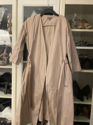 Beige trenchcoat från COS, strl 38 - Snygg beige trenchcoat från COS i storlek 38. Jackan har knytband i midjan, knappar och praktiska fickor. Den har en stilren och minimalistisk design med trekvartsärmar och är gjord i mjuk bomull. Perfekt för dig som gillar clean och enkel stil.