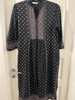Svart långklänning från Pragya - Svart långklänning med lila och vita detaljer från Pragya. Klänningen har ett geometriskt och småblommigt mönster, v-ringad hals och trekvartsärmar. Materialet känns lätt och mjukt, perfekt för dig som gillar boho-vibe och snygga mönster.