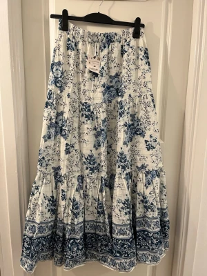Blommig långkjol från Zara M-L - Säljer en superfin långkjol från Zara i storlek M-L. Kjolen är vit med blått blommigt mönster och har volangdetaljer nertill. Materialet är mjuk bomull och midjan är elastisk för skön passform. Perfekt för vår och sommar!
