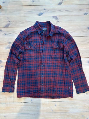 Rutig rödblå skjorta från Patagonia - Säljer en rutig skjorta från Patagonia i rött och mörkblått, tillverkad av ekologisk bomull. Skjortan har klassisk krage, två bröstfickor med knappar och långa ärmar med manschett. Perfekt för dig som gillar avslappnad stil och schyssta detaljer.
