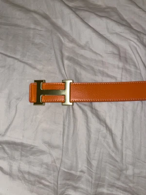 Svart och orange skinnbälte med H-spänne - Säljer ett snyggt vändbart bälte från H i svart och orange skinn med ett ikoniskt H-spänne i guld. Bältet har en grov textur och stilrena kanter, perfekt för att lyfta din outfit med lite lyxig vibe.
