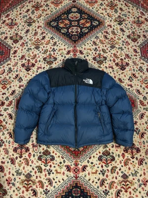 The North Face puffer - Blå north face puffer. Nypris 4000. Använd men bra skick. Små defekter men syns knappt. En på vänster arm nedtill och andra på ryggen nedtill, syns på priset. Den har fickor och luva som man kan väcka ut annars är den gömd. Storlek M. Mått: 52 cm axelbredd. 55cm bröstbredd. 67 cm längd. 55 cm armlängd