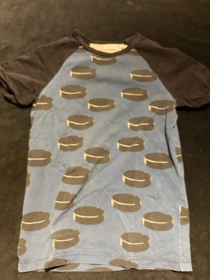 Blå t-shirt med svarta ärmar Sytt av Ami - Unik t-shirt med blå front och rygg, bruna korta ärmar och coolt mönster av bruna cirklar över hela plagget. Rund halsringning och mjukt bomullsmaterial gör den skön att bära. Perfekt för dig som gillar annorlunda prints och vill sticka ut.