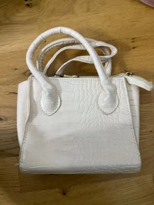 Vit handväska med krokopräglat mönster - Säljer en stilren vit handväska med krokopräglat mönster och två rejäla handtag. Väskan har en dragkedja upptill och en avtagbar axelrem. Perfekt för dig som vill ha en chic accessoar med lite edge. Materialet är syntet och ytan har en glansig finish.