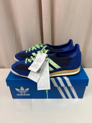 Adidas sneakers blå och mint - Snygga Adidas sneakers i marinblått och mintgrönt med klassiska tre ränder på sidan. Ovandelen är i mocka och textil, med mintgröna snören och detaljer i orange på mellansulan. Yttersulan är svart och har ett mönstrat grepp. Perfekt för dig som gillar retrostil och färg.