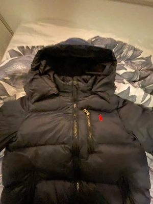 Marinblå pufferjacka från Polo Ralph Lauren - Marinblå pufferjacka från Polo Ralph Lauren med huva och röd logga på bröstet. Jackan har flera fickor med dragkedja och en clean, quiltad design. Perfekt för kalla dagar och har en riktigt schysst streetstil. Materialet är polyester och dun.