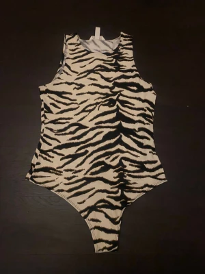 Zebramönstrad ärmlös bodysuit - Säljer en cool ärmlös bodysuit med zebramönster i svart och beige. Toppen har rund hals och är figurnära med högt skurna ben. Perfekt för att sticka ut med ett djuriskt print. Materialet känns mjukt och stretchigt, troligen polyester eller syntet.