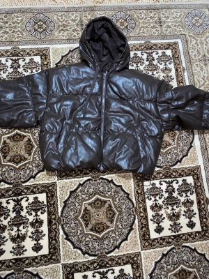 Brun pufferjacka med huva - Säljer en brun pufferjacka med huva och dragkedja framtill. Jackan har ett glansigt yttre i syntetmaterial som liknar skinn, ribbade muddar vid ärmslut och snörning i nederkant. Perfekt för dig som gillar streetstyle och vill hålla dig varm.