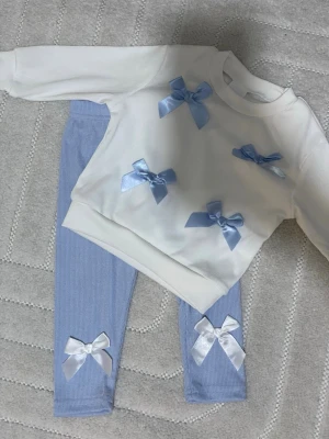 Blå leggings med rosetter - Säljer ett par ljusblå leggings med ribbad struktur och två vita satinsrosetter längst ner på benen. Leggingsen är mjuka och stretchiga, perfekta för en chill och söt look. Passar dig som gillar detaljer och vill ha något extra i vardagen.