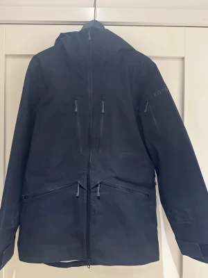 Svart skidjacka från EQPE  - Svart skidjacka från EQPE med flera vattentäta dragkedjor, justerbar huva och praktiska fickor på bröstet och ärmen. Jackan är en skaljacka med recco (lavinutrustning)och perfekt för skidbacken. Är sparsamt använd utan några defekter. Nypris 2999 