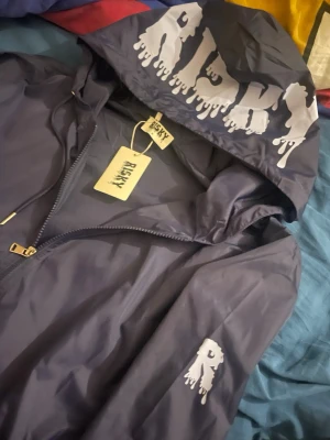 Mörkblå vindjacka från Risky - Snygg mörkblå vindjacka från Risky med stor ljusblå logga på huvan och mindre tryck på ärmen. Jackan har dragkedja, huva med snörning och är tillverkad i polyester. Perfekt för dig som gillar streetwear och vill sticka ut med coola detaljer. Bara testad hemma, aldrig direkt använd. Nypris 849kr.