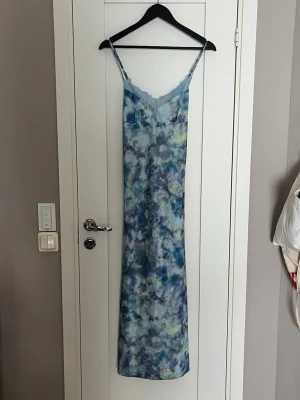 Lång blåmönstrad slipklänning med spets - Säljer en lång slipklänning med tunna axelband och vacker spetskant upptill. Klänningen har ett blått, lila och grönt abstrakt mönster och är gjord i ett glansigt, mjukt material som känns som satin eller polyester. Perfekt för dig som gillar en chill men ändå snygg vibe.