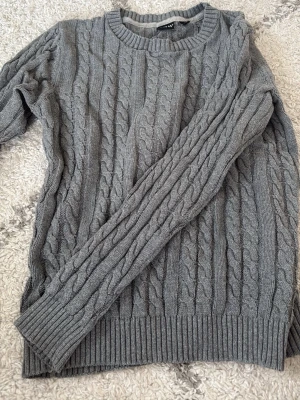 Grå stickad tröja från CRW - Säljer en grå stickad tröja från CRW i storlek XS. Tröjan har ett klassiskt flätmönster och ribbade muddar vid hals, ärmslut och nederkant. Perfekt för kyliga dagar och enkel att matcha med jeans eller kjol.