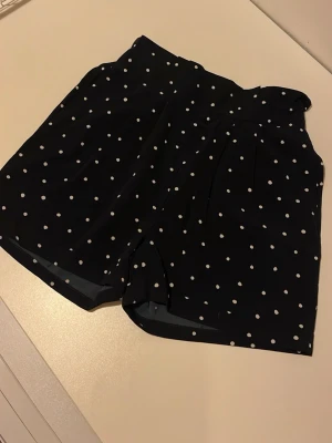 Prickiga shorts från Name It - Gulliga marinblå shorts med vita prickar från Name It, storlek 164. De har en högmidjad med elastisk midja och är gjorda i ett mjukt och lätt bomullsmaterial. Perfekta för varma dager med loose fit! Aldrig använda!