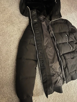 Svart pufferjacka Supply & Demand S - Säljer en svart pufferjacka från Supply & Demand i storlek S. Jackan har huva med mjuk fuskpäls på insidan, silverfärgade dragkedjor och flera fickor framtill. Oversized känsla och ribbade muddar vid ärmar och midja. Perfekt för kalla dagar.