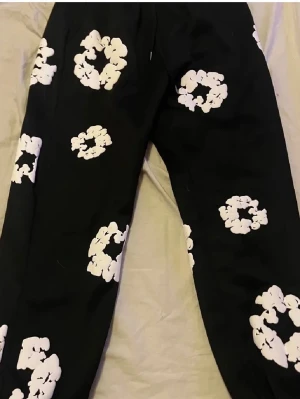 Denim Tears mjukisbyxor - Svarta byxor med stora vita blommiga mönster över hela benen. Byxorna har en avslappnad passform och är perfekta för dig som vill sticka ut med en unik stil. Materialet känns mjukt och bekvämt mot huden. Snygga att matcha med en enkel t-shirt eller hoodie.