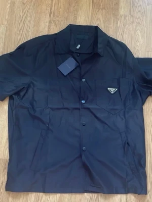 Svart overshirt från Prada - Säljer en svart overshirt från Prada med klassisk krage och korta ärmar. Skjortan har knappar framtill och en bröstficka med Pradas ikoniska metalltriangel. Perfekt för dig som gillar stilrena designer och exklusiva märken.