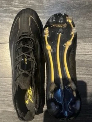 Svarta adidas F50 fotbollsskor elite storlek 44/2 - Säljer ett par svarta adidas F50 fotbollsskor med gula detaljer och snörning. Skorna har en låg profil och är tillverkade i syntetmaterial med mönstrad ovansida och platt sula med dobbar för bra grepp på planen. Perfekt för dig som vill ha snabba och lätta skor.