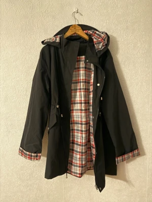 Svart parkajacka med rutigt foder - Svart parkajacka med huva och rutigt foder i vitt, rött och grått. Jackan har dragsko i midjan, långa ärmar och detaljer i samma rutiga tyg vid ärmslut och huva. Stängs med dragkedja och tryckknappar. Perfekt för kyliga dagar.