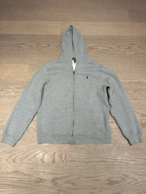 Grå hoodie från Polo Ralph Lauren XL - Klassisk grå hoodie från Polo Ralph Lauren i storlek 170. Tröjan har dragkedja, huva och två fickor framtill. Tillverkad i mjuk bomullsmix och har den ikoniska lilla loggan broderad på bröstet. Perfekt för chill dagar eller när du vill ha en clean look. Frakt ingår ej. Skriv ifall ni vill ha andra bilder.