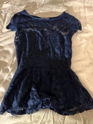 Spetsblus🦕🌟 - Säljer en mörkblå blus från Gina Tricot i storlek XS. Toppen är helt täckt av vacker spets med blommönster och har korta ärmar samt rund halsringning. Den har en lätt utsvängd peplum nedtill och är perfekt för dig som gillar feminina detaljer💙🌟.