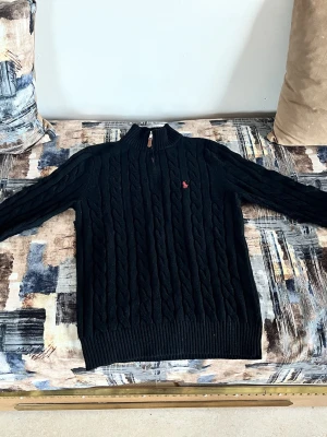 Svart stickad tröja Polo Ralph Lauren - Kabelstickad svart tröja från Polo Ralph Lauren med röd broderad logga på bröstet. Tröjan har hög krage med half zip-dragkedja och ribbade muddar. Perfekt för kyliga dagar och riktigt snygg till jeans eller chinos.