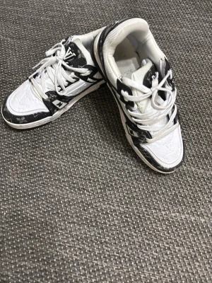 Louis Vuitton vita och svarta sneakers - Säljer ett par riktigt snygga Louis Vuitton sneakers i vitt och svart med klassiskt LV-monogram. Skorna har snörning, rund tå och platt sula. Materialmix av skinn och textil med detaljer i svart och vitt samt monogrammönster på flera partier. Perfekta för dig som vill sticka ut.