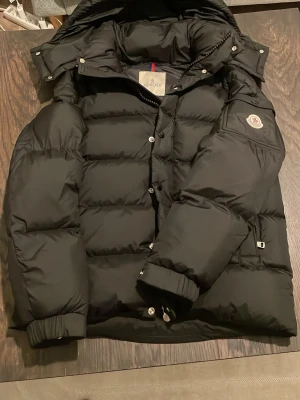 Svart Moncler vezere - Svart pufferjacka från Moncler med huva och ikonisk logga på ärmen. Jackan har quiltad design, dragkedja och knappar framtill samt ribbade muddar vid ärmslut. Insidan har ett coolt serietryck och innerficka med dragkedja. Perfekt för kalla dagar.
